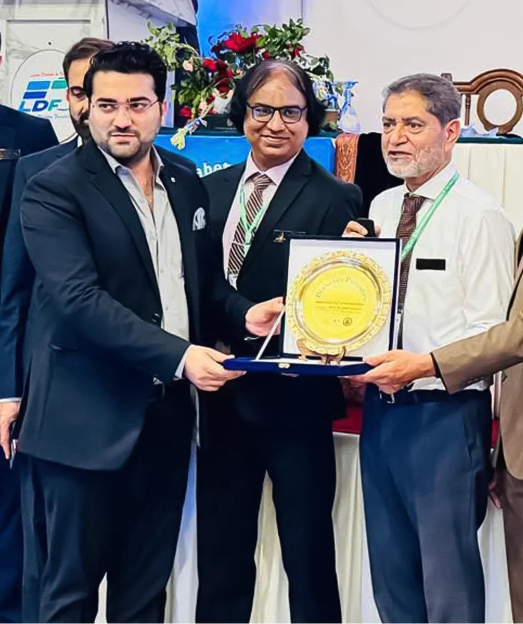 Men in formal attire presenting and receiving a gold award plaque at a ceremony